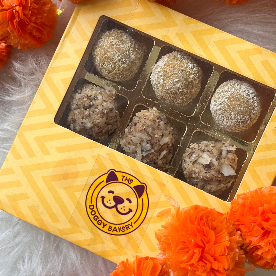 A box of assorted Ladoos, which are traditional Indian sweets, presented in a decorative box with The Doggy Bakery logo, surrounded by orange flowers.