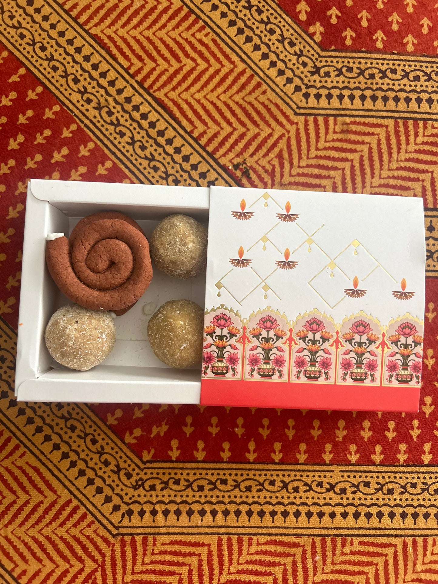 A box of assorted festive treats, including what appears to be besan laddoos and possibly cheese crackers, presented on a red and patterned surface.