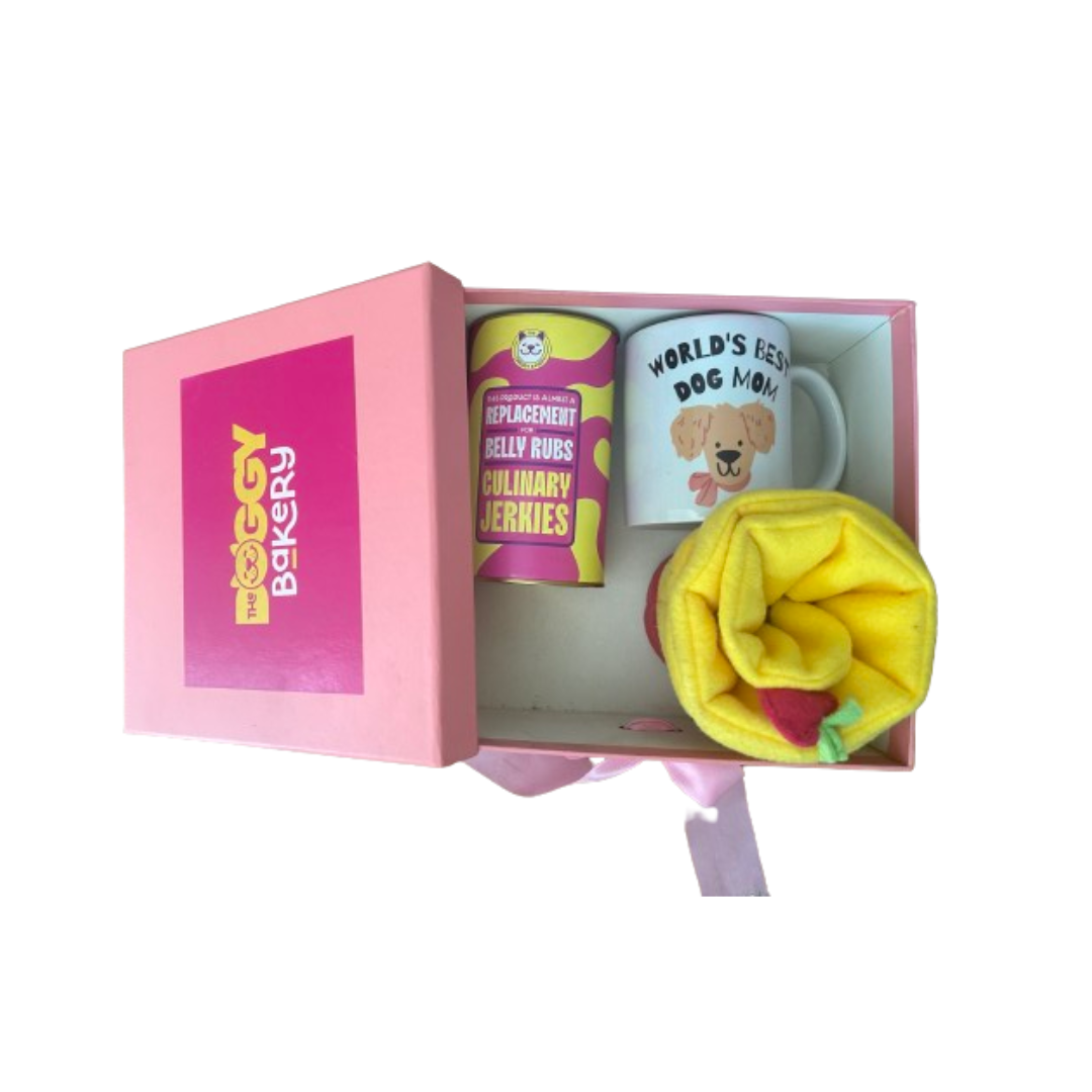 A gift box with a 'Dog Mom' theme, containing a coffee mug, a yellow flower, and a stuffed toy, all presented on a pink background with 'The Doggy Bakery' branding.