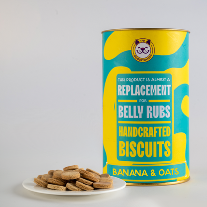 A canister labeled 'This Product is a Replacement for Belly Rubs Handcrafted Biscuits' with a graphic of banana slices and oats, indicating the product is a pet treat made with banana and oats. A plate with a few dog biscuits is also visible in front of the canister.